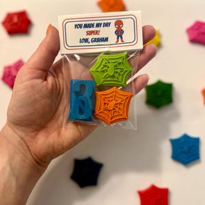 May include: A clear plastic bag contains a blue number 3 crayon, and two spiderweb-shaped crayons, one green and one orange. The bag has a label with the text "YOU MADE MY DAY SUPER! LOVE, GRAHAM" and a cartoon superhero.