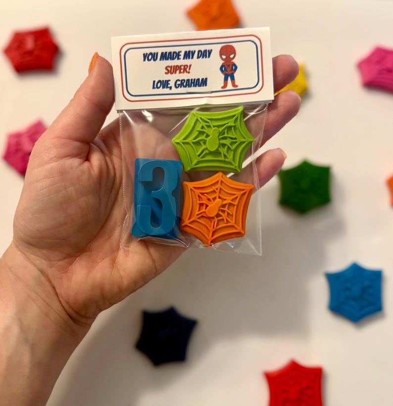 May include: A clear plastic bag contains a blue number 3 crayon, and two spiderweb-shaped crayons, one green and one orange. The bag has a label with the text "YOU MADE MY DAY SUPER! LOVE, GRAHAM" and a cartoon superhero.