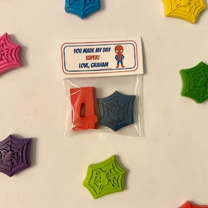 May include: A clear plastic bag contains a red number 4 and a blue spiderweb-shaped item. The bag is attached to a card with the text "YOU MADE MY DAY SUPER! LOVE, GRAHAM" and a Spider-Man graphic. Scattered around are colorful spiderweb-shaped items.