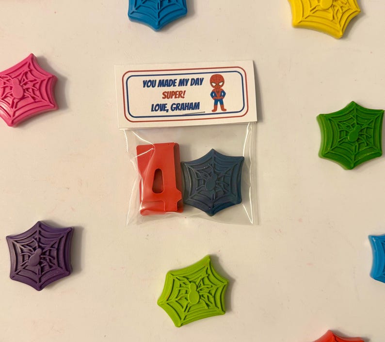 May include: A clear plastic bag contains a red number 4 and a blue spiderweb-shaped item. The bag is attached to a card with the text "YOU MADE MY DAY SUPER! LOVE, GRAHAM" and a Spider-Man graphic. Scattered around are colorful spiderweb-shaped items.