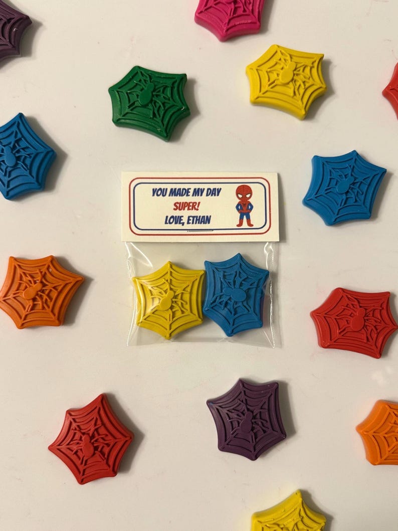 May include: A collection of colorful, spider web-shaped crayons in various colors, including yellow, blue, red, and purple. A small package contains two yellow and blue crayons. A card reads "YOU MADE MY DAY SUPER! LOVE, ETHAN" with a Spider-Man graphic.