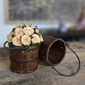 May include: A rustic wooden bucket filled with pale pink roses and green leaves, with a second bucket lying on its side. The buckets have metal handles and are set on a wooden surface, creating a charming floral arrangement.