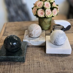 Peut inclure: Trois sphères décoratives sur des socles en marbre. Une sphère est vert foncé, une autre rose clair et la troisième blanche avec des veines grises. Un bouquet de roses roses dans un vase vert est en arrière-plan.