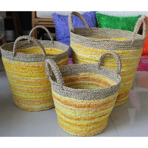 May include: Three woven storage baskets in varying sizes. The baskets are cylindrical with sturdy handles. The main body of each basket features horizontal stripes of yellow and orange hues, while the top and handles are a neutral brown.