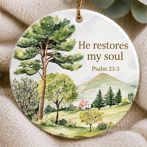 May include: A round white ceramic ornament featuring a watercolour landscape illustration of a green pine tree, a small house with a red roof, and rolling hills. The text "He restores my soul" and "Psalm 23-3" is printed in brown lettering.