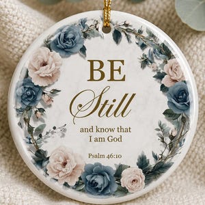 May include: A round white ceramic ornament featuring a gold-colored floral wreath of blue and pale pink roses. The center text reads: BE Still and know that I am God Psalm 46:10. The ornament hangs from a gold string against a textured fabric background.