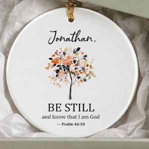 May include: A round white ceramic ornament featuring a colorful tree illustration, the name Jonathan, and the text BE STILL and know that I am God — Psalm 46:10. The ornament hangs inside a sage green gift box with a silver ribbon.