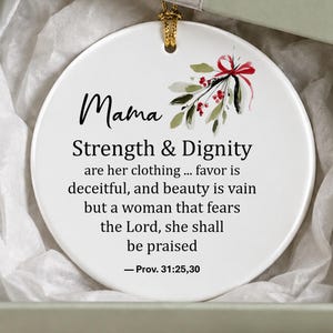 Può includere: Un ornamento rotondo in ceramica bianca con un rametto di vischio con bacche rosse e testo nero che recita: Mama, Strength & Dignity are her clothing... favor is deceitful, and beauty is vain but a woman that fears the Lord, she shall be praised - Prov. 31:25,30.