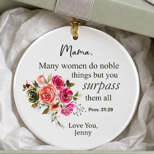 May include: A round white ceramic ornament featuring a floral illustration and the text: Mama, Many women do noble things but you surpass them all Prov. 31:29 Love You, Jenny. The ornament is inside a sage green gift box with a silver ribbon.