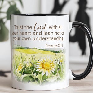 May include: White ceramic mug with a black handle and rim. The mug features a watercolor design of sunflowers in a field with the text "Trust the Lord with all your heart and lean not on your own understanding Proverbs 3:5-6."