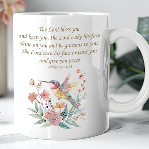 Puede incluir: Taza de cerámica blanca con un colibrí colorido y un diseño floral. La taza tiene un asa curva e incluye el texto "The Lord bless you...and give you peace" de Filipenses 4:13.