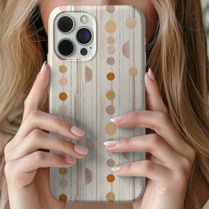 Op de afbeelding: Een beige telefoonhoesje met een geometrisch patroon van verticale lijnen, halve cirkels en stippen in bruin- en oranje tinten. De hoes heeft een camera-uitsparing en wordt voor het gezicht van een persoon gehouden.