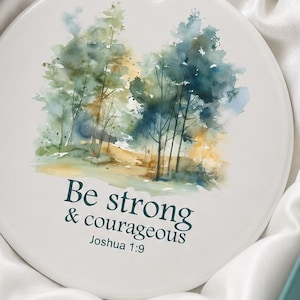 May include: A round white ceramic ornament featuring a watercolor forest illustration in shades of green, blue, and yellow. The text reads "Be strong & courageous Joshua 1:9" in dark blue. It includes a gold hanging cord and a silver ribbon.