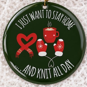 May include: A round dark green ceramic ornament featuring a red heart-shaped yarn ball, a red mug with a smiling face, and two red mittens with white snowflake patterns. The text reads I JUST WANT TO STAY HOME AND KNIT ALL DAY in white.