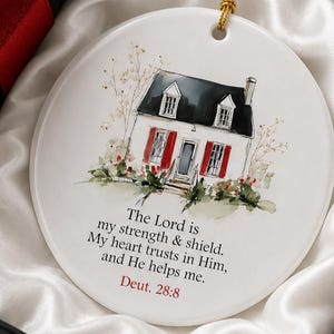 May include: A round white ceramic ornament featuring a watercolor illustration of a house with red shutters and black roof. Below the house, the text reads: The Lord is my strength & shield. My heart trusts in Him, and He helps me. Deut. 28:8.