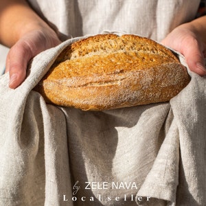 May include: A person holding a freshly baked loaf of bread wrapped in a linen cloth. The bread is golden brown and has a crusty exterior. The text "by ZELE NAVA Local seller" is visible at the bottom of the image.