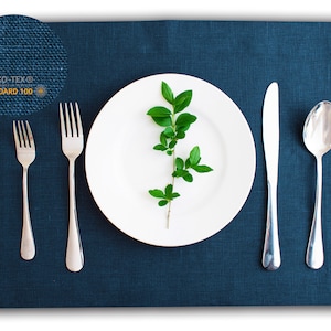 May include: A white plate with a sprig of green leaves sits on a dark blue placemat. A fork, knife, and spoon are arranged to the right of the plate. The placemat has a label that reads "OEKO-TEX® STANDARD 100."