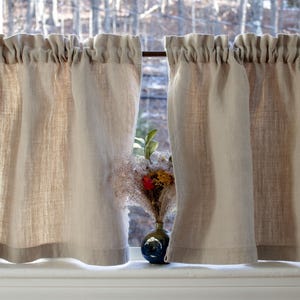 May include: A pair of beige linen cafe curtains with a ruffled top, hanging in a window with a small vase of dried flowers in the center.