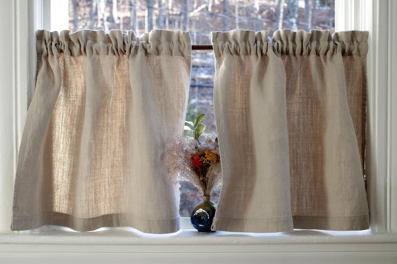 May include: A pair of beige linen cafe curtains with a ruffled top, hanging in a window with a small vase of dried flowers in the center.