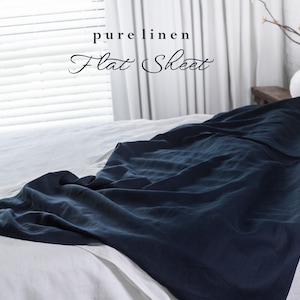 May include: A dark blue linen flat sheet is folded and draped over a white bed. The sheet is made of 100% pure linen and is soft and breathable. The text "pure linen Flat Sheet" is written in white on a white background.