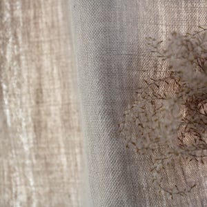 May include: Close-up of a natural linen curtain with a textured weave. The curtain is a light beige color and has a soft, drapey feel.