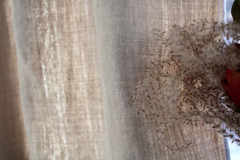 May include: Close-up of a natural linen curtain with a textured weave. The curtain is a light beige color and has a soft, drapey feel.