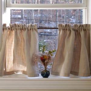 May include: A pair of beige and brown striped linen valance curtains hang in a window with a view of trees and a small bouquet of flowers in a blue vase on the windowsill.