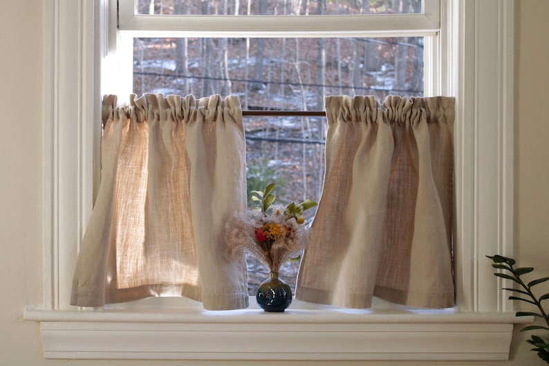May include: A pair of beige and brown striped linen valance curtains hang in a window with a view of trees and a small bouquet of flowers in a blue vase on the windowsill.