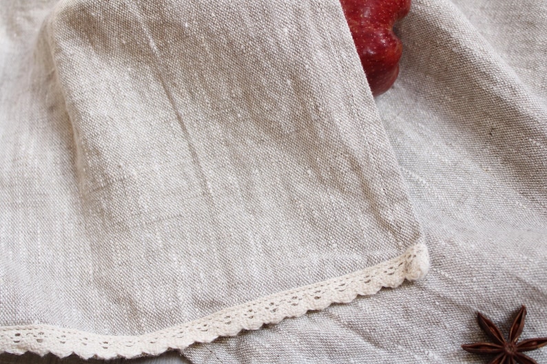 May include: A close-up of a beige linen fabric with a white lace trim. The fabric has a subtle texture and a slightly rough feel.