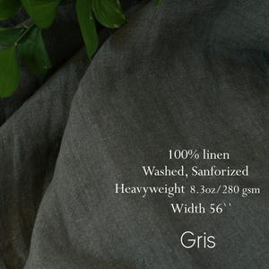 Pu&ograve; includere: Un tessuto di lino grigio scuro con una trama visibile. Il testo sul tessuto recita: 100% linen, Washed, Sanforized, Heavyweight 8.3oz/280 gsm, Width 56'', Gris. Il materiale &egrave; drappeggiato in morbide pieghe con foglie verdi in alto.