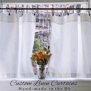May include: White linen curtains hanging in a window, with a floral arrangement in a glass jar on the windowsill. The curtains have a neutral-colored border. Text on the windowsill reads "Custom Linen Curtains Hand-made in the US."