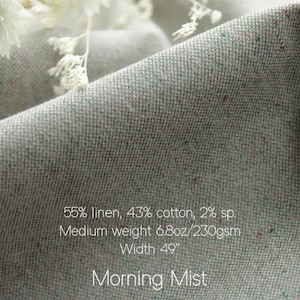 May include: A light gray fabric with subtle multicolored flecks, featuring the text: 55% linen, 43% cotton, 2% sp. Medium weight 6.8oz/230gsm Width 49". Morning Mist. The material has a textured weave and is draped in soft folds.