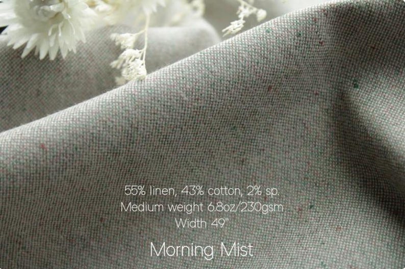 May include: A light gray fabric with subtle multicolored flecks, featuring the text: 55% linen, 43% cotton, 2% sp. Medium weight 6.8oz/230gsm Width 49". Morning Mist. The material has a textured weave and is draped in soft folds.