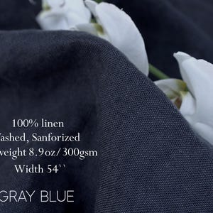 Pu&ograve; includere: Un pezzo di tessuto in lino 100% grigio blu con finitura lavata e sanforizzata. Il tessuto &egrave; pesante con 300 g/m&sup2; e ha una larghezza di 54 pollici. Fiori di orchidea bianchi sono posizionati lungo il bordo piegato del tessuto scuro.