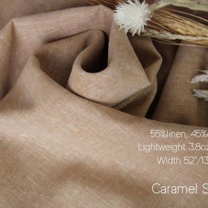 May include: A piece of caramel sand colored fabric with a blend of 55% linen and 45% cotton. The material is lightweight at 3.8oz and has a width of 52 inches. Dried wheat stalks and a white flower are placed on the folded fabric surface.
