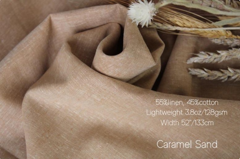 May include: A piece of caramel sand colored fabric with a blend of 55% linen and 45% cotton. The material is lightweight at 3.8oz and has a width of 52 inches. Dried wheat stalks and a white flower are placed on the folded fabric surface.