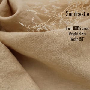 Pu&ograve; includere: Un tessuto beige con una trama visibile, etichettato Sandcastle, Irish 100% Linen, Weight 193g, Width 147cm. Un delicato rametto di pianta bianca essiccata poggia sulla superficie piegata e drappeggiata del materiale tessile color marrone chiaro.