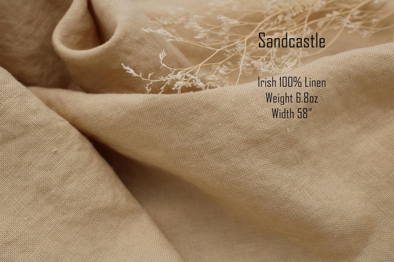 Pu&ograve; includere: Un tessuto beige con una trama visibile, etichettato Sandcastle, Irish 100% Linen, Weight 193g, Width 147cm. Un delicato rametto di pianta bianca essiccata poggia sulla superficie piegata e drappeggiata del materiale tessile color marrone chiaro.