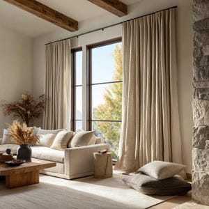 May include: A living room with neutral tones, featuring beige curtains, a light-colored sofa, and a wooden coffee table. The room is illuminated by natural light from a large window, with decorative pillows and a rug.