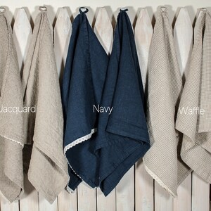 May include: Three linen kitchen towels hanging on a white picket fence. The towels are in different weaves: jacquard, waffle, and plain. The towels are beige, navy blue, and beige.