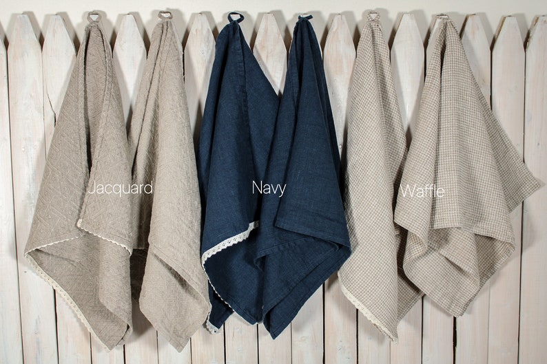 May include: Three linen kitchen towels hanging on a white picket fence. The towels are in different weaves: jacquard, waffle, and plain. The towels are beige, navy blue, and beige.