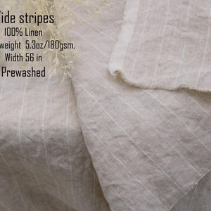 May include: A piece of prewashed 100% linen fabric featuring wide stripes in a natural beige tone. The textile has a medium weight of 5.3oz and a width of 56 inches. The fabric displays a visible woven texture with raw, frayed edges.