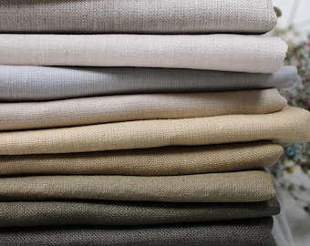 May include: A pile of folded linen fabric in a range of neutral colours, including white, beige, grey, and black. The fabrics have a textured appearance, suggesting a natural fibre. The image is well-lit, highlighting the different shades and textures.