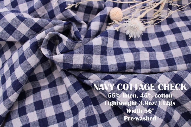 May include: Navy blue and white gingham check fabric with a soft, draped texture. Text on the fabric reads: NAVY COTTAGE CHECK, 55% linen, 45% cotton, Lightweight 3.9oz/132gs, Width 56", Pre-washed. A small cluster of dried white flowers rests on top.