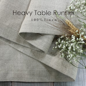 May include: A heavy linen table runner in a neutral beige color. The text "Heavy Table Runner 100% linen" is visible. The runner is folded, revealing its texture. Wheat stalks and white flowers are arranged on the side.
