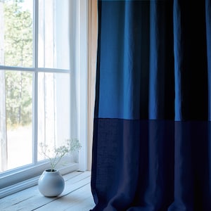 May include: A blue linen curtain with a darker blue bottom panel. The curtain is hanging in a window with a white frame. A small white vase with white flowers sits on the windowsill.