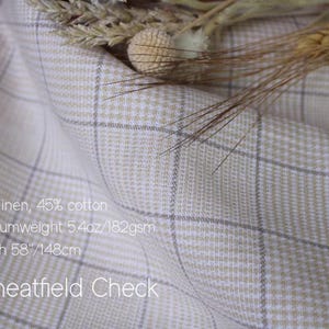 May include: A folded piece of fabric with a beige and white check pattern, labeled Wheatfield Check. The text states 55% linen, 45% cotton, mediumweight 5.4oz, and a width of 58 inches. Dried wheat stalks are resting on the top edge of the fabric.