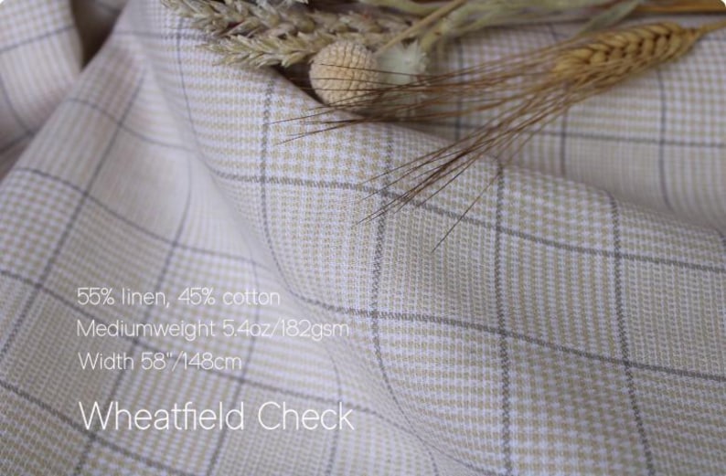 May include: A folded piece of fabric with a beige and white check pattern, labeled Wheatfield Check. The text states 55% linen, 45% cotton, mediumweight 5.4oz, and a width of 58 inches. Dried wheat stalks are resting on the top edge of the fabric.