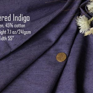 May include: A piece of heathered indigo fabric with a visible weave, featuring text that reads: Heathered Indigo, 55% linen, 45% cotton, Medium-weight 7.1 oz/241gsm, Width 55". A copper-colored coin rests on the fabric, and dried white flowers are visible.