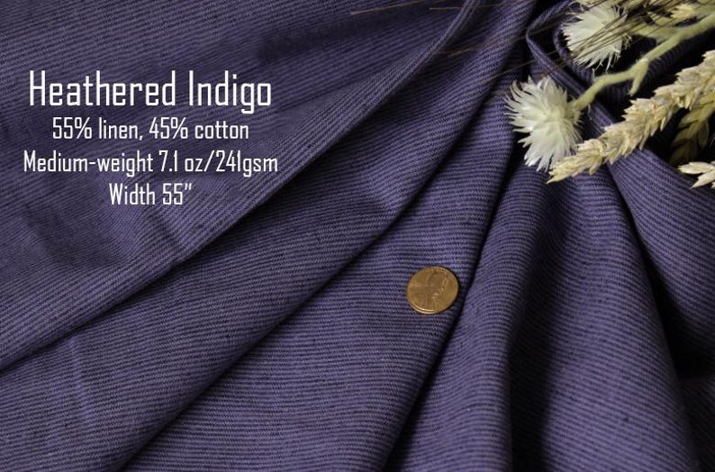 May include: A piece of heathered indigo fabric with a visible weave, featuring text that reads: Heathered Indigo, 55% linen, 45% cotton, Medium-weight 7.1 oz/241gsm, Width 55". A copper-colored coin rests on the fabric, and dried white flowers are visible.
